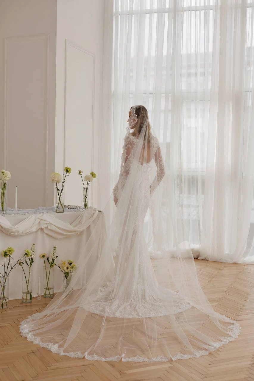 Bride wearing a fitted lace wedding dress with long sleeves and a cathedral-length veil in a bright luxury bridal setting.
