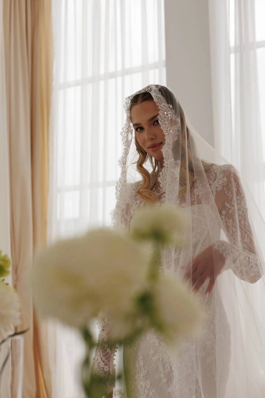 Bride wearing a lace wedding gown with sheer sleeves and lace cathedral veil near large windows.