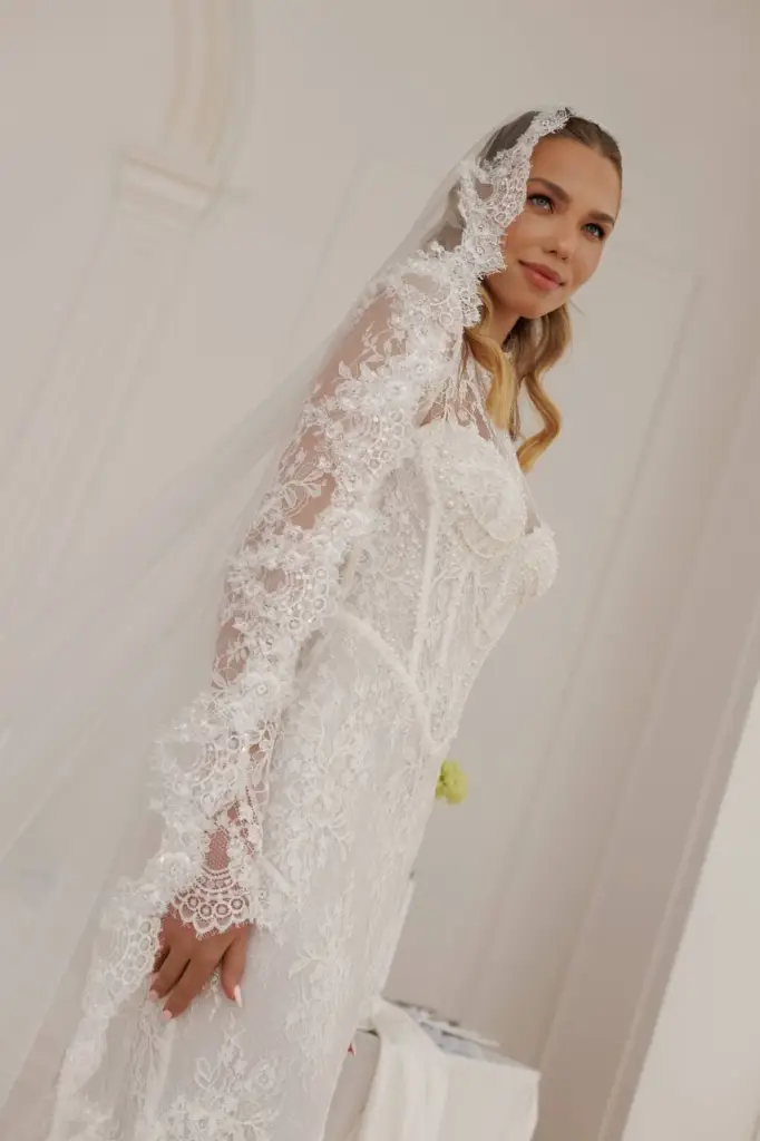 Bride modeling a fitted lace wedding gown with embroidered sleeves and lace veil.